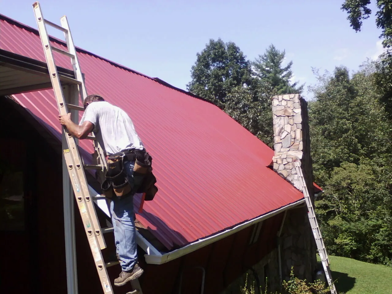 Roofer on a ladder beside a red metal roof and stone chimney — Soffit & Fascia Repair by Roofing Brothers in Spring Lake